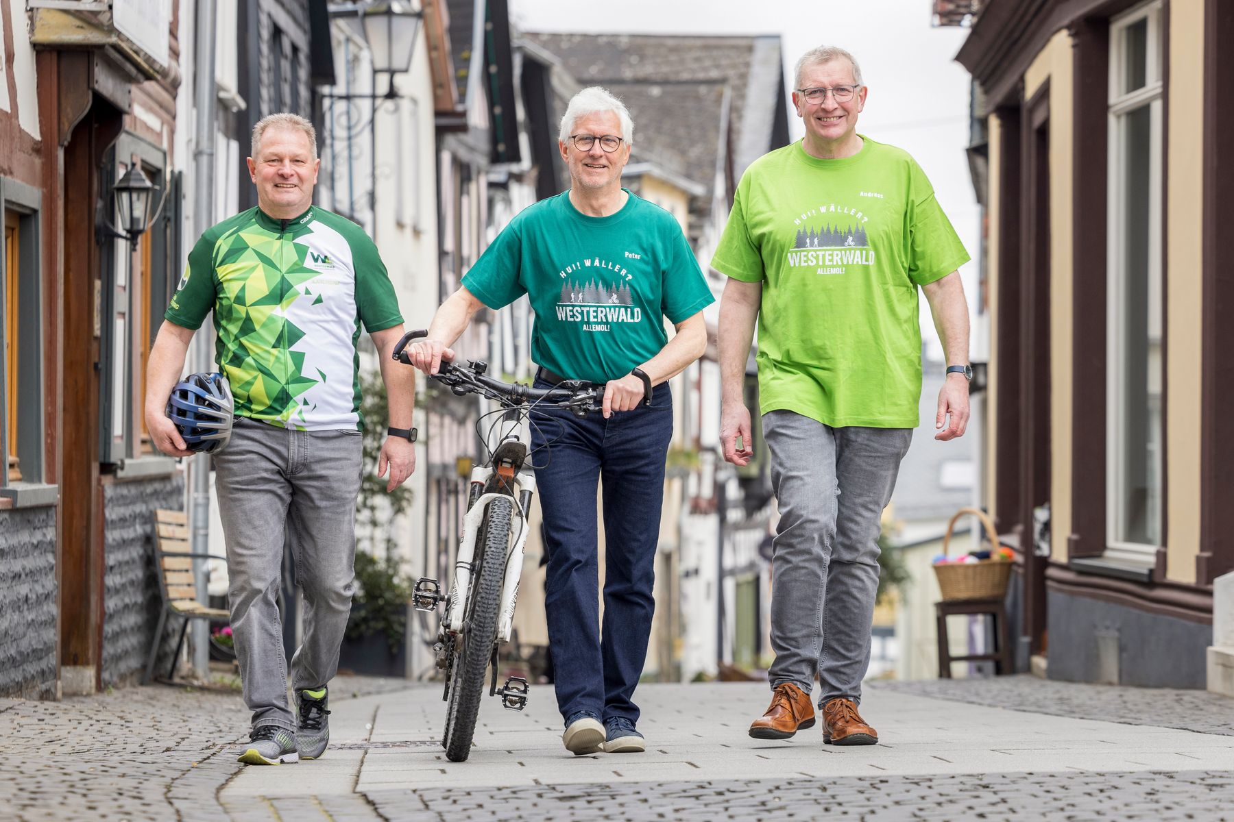 Drei lächelnde Männer in Freizeitkleidung gehen eine Kopfsteinpflasterstraße entlang; zwei gehen, während der Mann in der Mitte ein Fahrrad schiebt. Alle drei tragen grüne "Westerwald"-Hemden, und hinter ihnen säumen traditionelle Gebäude die Straße.